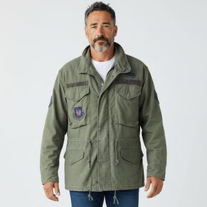 May include: An olive green field jacket with a high collar, four front pockets, and a drawstring waist. The jacket features various military-style patches, including one labelled SADOWSKI, worn over a white t-shirt, dark blue jeans, and white trainers.