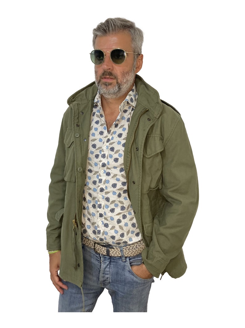 May include: A man wearing a green field jacket over a white button-down shirt with a blue and brown leaf pattern. He is wearing blue jeans and a brown woven belt. He is wearing sunglasses.