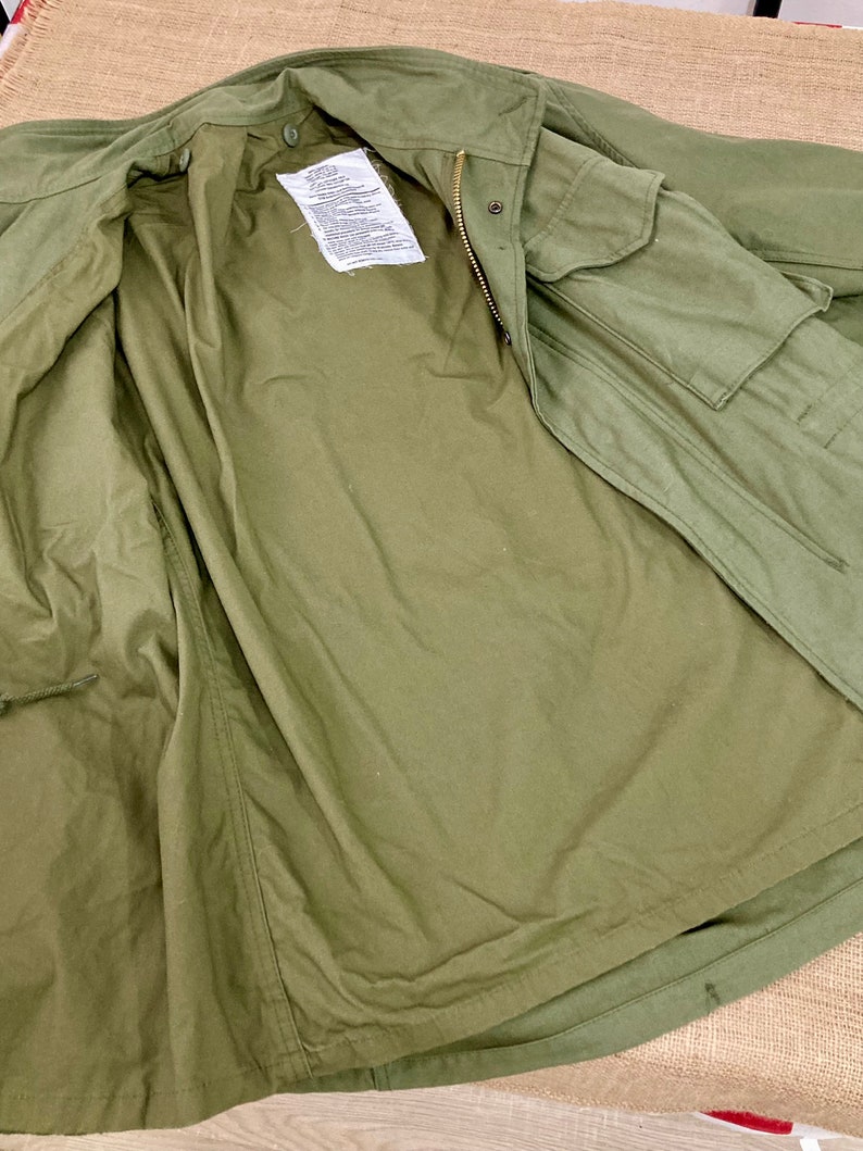 May include: A green military-style jacket with a zipper and a flap pocket. The jacket is unbuttoned and turned inside out, showing the lining.