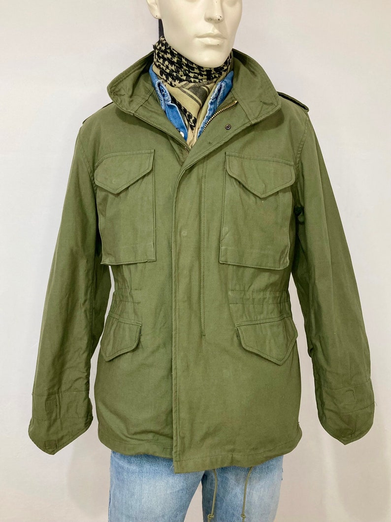 May include: A green military-style jacket with a zipper closure and multiple pockets. The jacket has a drawstring waist and a hood.