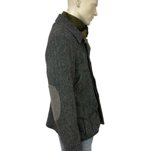 May include: A gray herringbone tweed jacket with elbow patches, worn over a green scarf and blue jeans. The jacket has a button closure and a classic design, suitable for cooler weather.