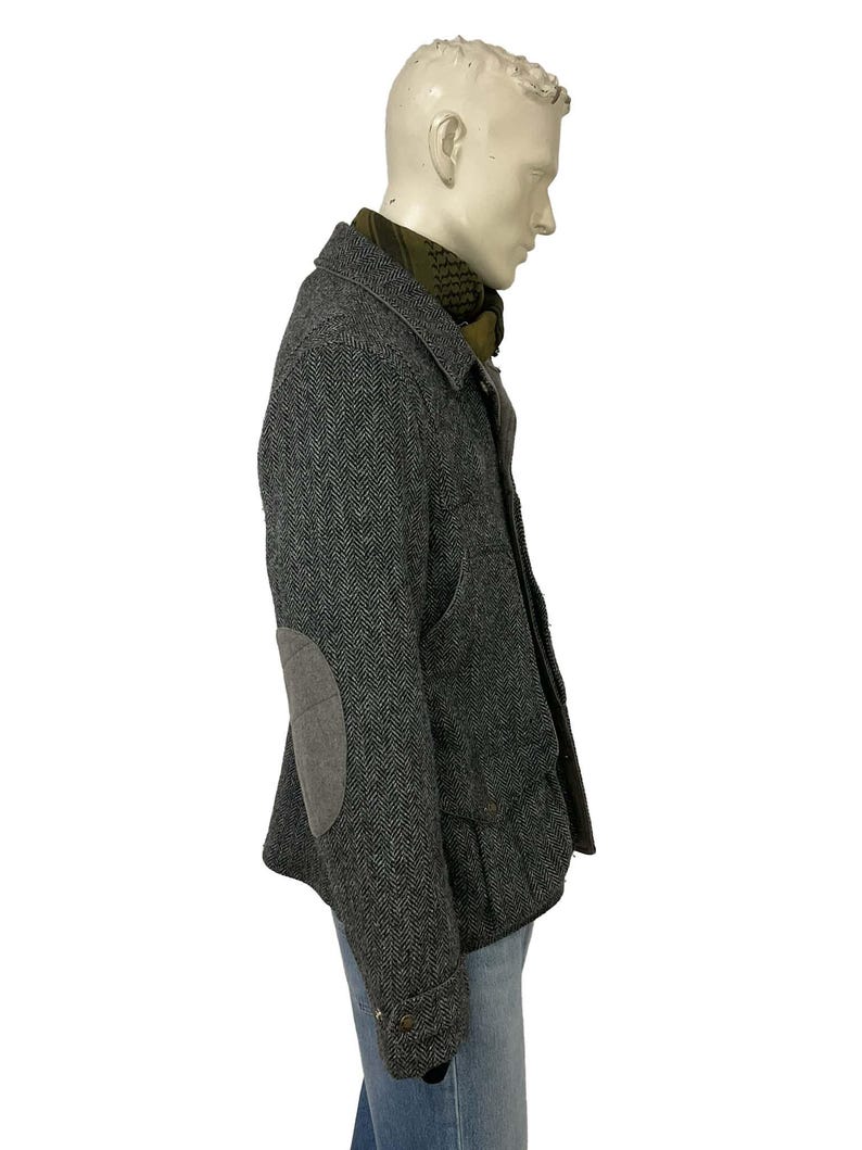 May include: A gray herringbone tweed jacket with elbow patches, worn over a green scarf and blue jeans. The jacket has a button closure and a classic design, suitable for cooler weather.