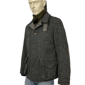 May include: A gray herringbone tweed jacket with a notched collar, two front pockets, and elbow patches. The jacket has a button closure and is worn over a green and black patterned scarf. The mannequin is also wearing blue jeans.