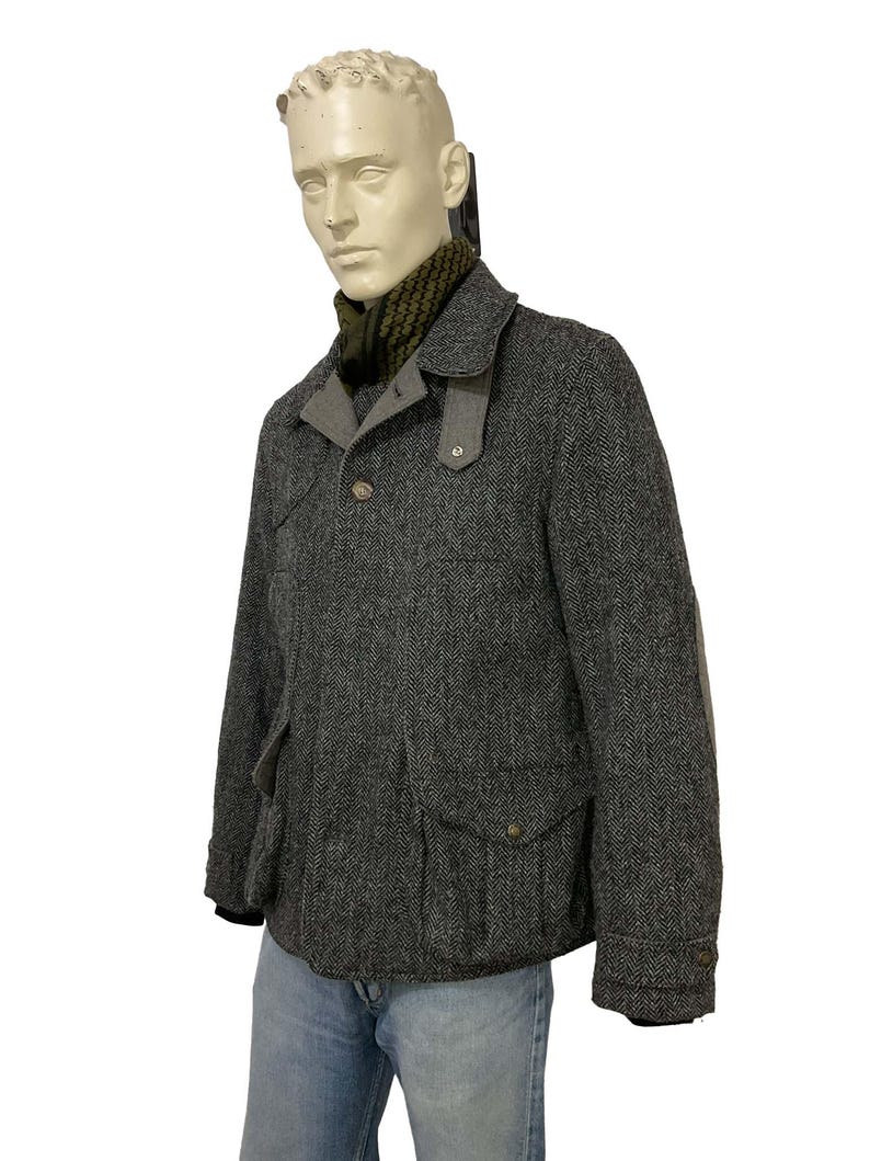 May include: A gray herringbone tweed jacket with a notched collar, two front pockets, and elbow patches. The jacket has a button closure and is worn over a green and black patterned scarf. The mannequin is also wearing blue jeans.