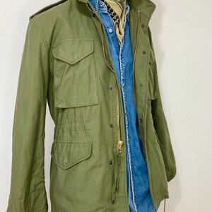 May include: A green military-style jacket with a zipper closure and multiple pockets. The jacket is worn over a blue denim shirt and a patterned scarf.