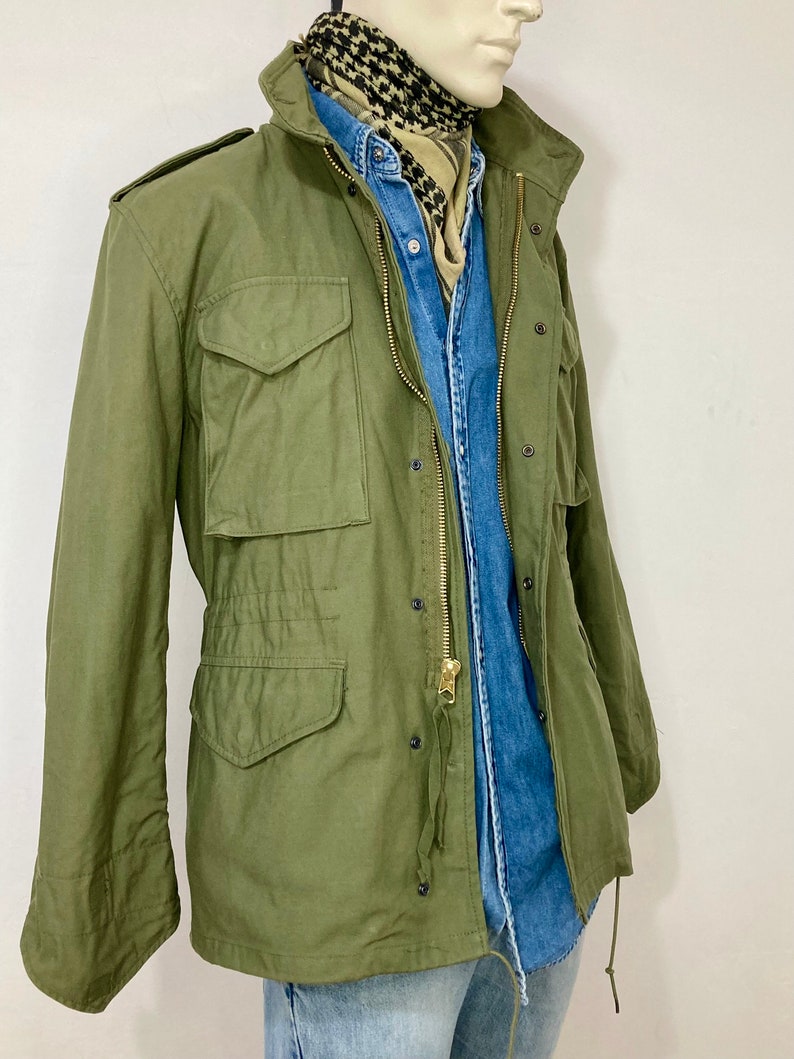 May include: A green military-style jacket with a zipper closure and multiple pockets. The jacket is worn over a blue denim shirt and a patterned scarf.