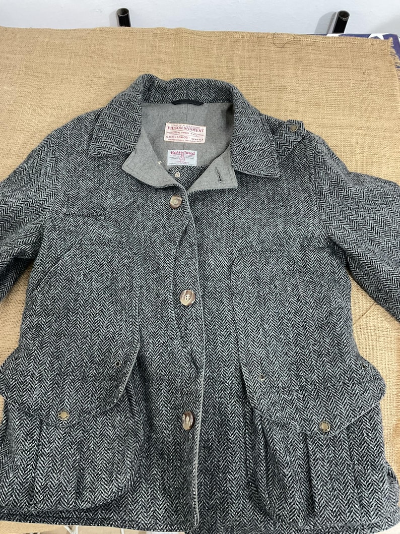 May include: A gray herringbone tweed jacket with a notched collar, button closures, and two front pockets. The jacket has a gray lining and a label that reads "Filson Garment".