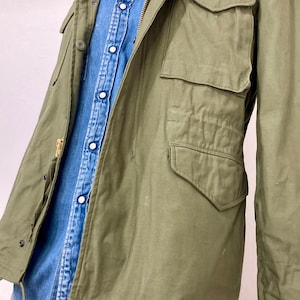 May include: A green military-style jacket with a zipper closure and multiple pockets. The jacket is worn over a blue denim shirt.