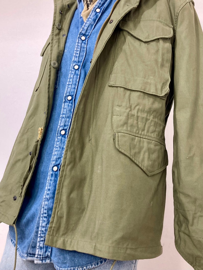May include: A green military-style jacket with a zipper closure and multiple pockets. The jacket is worn over a blue denim shirt.