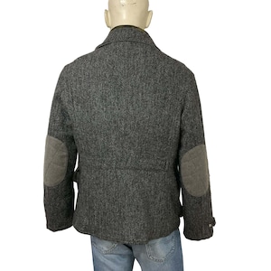 May include: A gray herringbone tweed jacket with gray elbow patches. The jacket has a classic design with a notched collar and buttoned cuffs. The jacket is displayed on a mannequin.