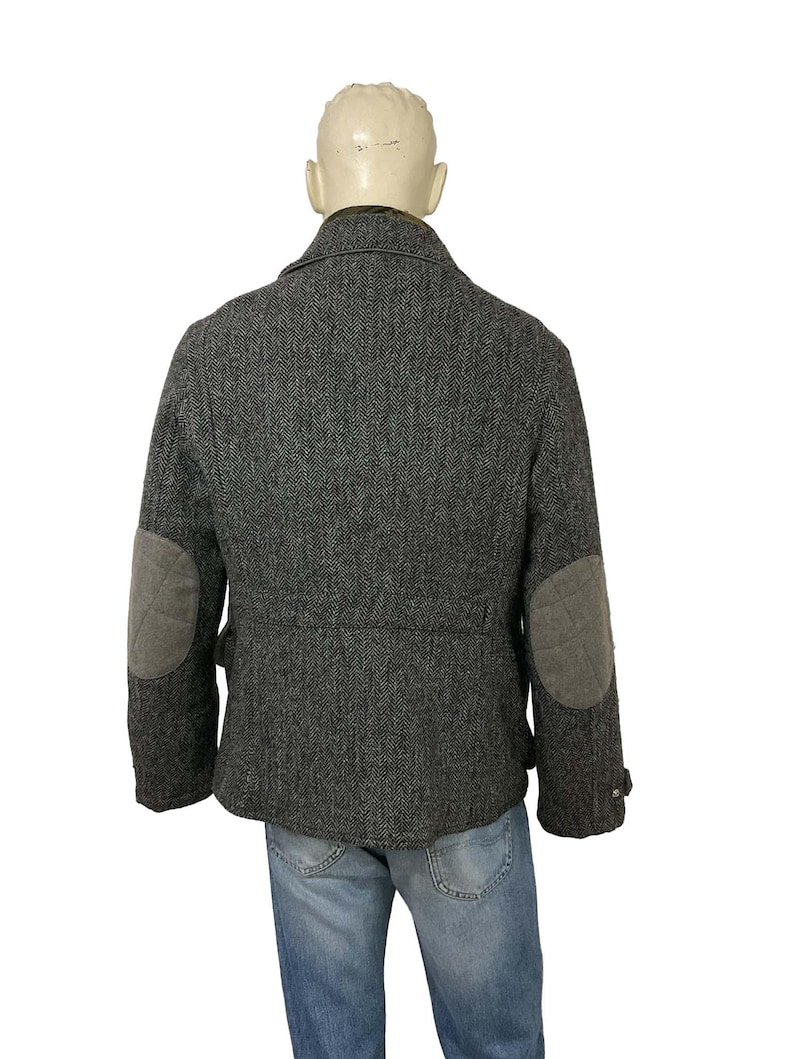 May include: A gray herringbone tweed jacket with gray elbow patches. The jacket has a classic design with a notched collar and buttoned cuffs. The jacket is displayed on a mannequin.