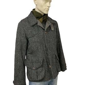 May include: A gray herringbone tweed jacket with a notched collar, two front flap pockets, and button closures. The jacket is worn over a gray shirt and paired with a green patterned scarf and blue jeans. The jacket has a classic, tailored look.