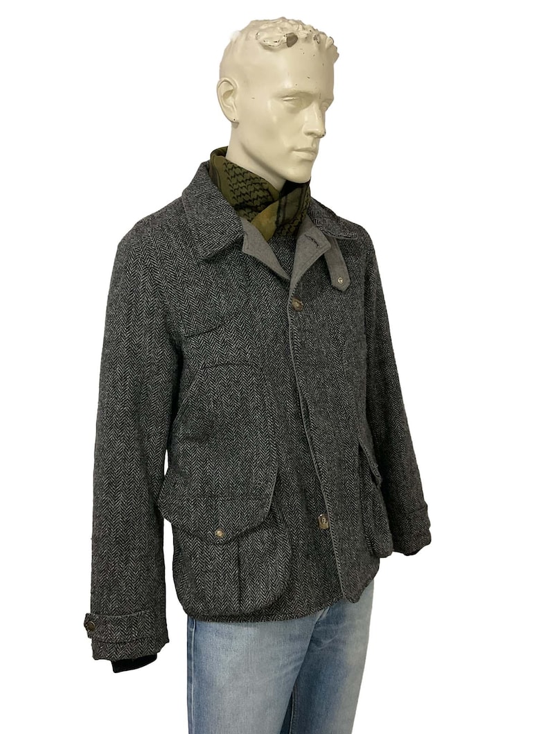 May include: A gray herringbone tweed jacket with a notched collar, two front flap pockets, and button closures. The jacket is worn over a gray shirt and paired with a green patterned scarf and blue jeans. The jacket has a classic, tailored look.