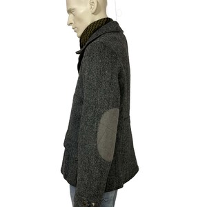 May include: A gray herringbone tweed jacket with elbow patches. The jacket has a notched collar, button closures, and a dark green scarf. The jacket is displayed on a mannequin.