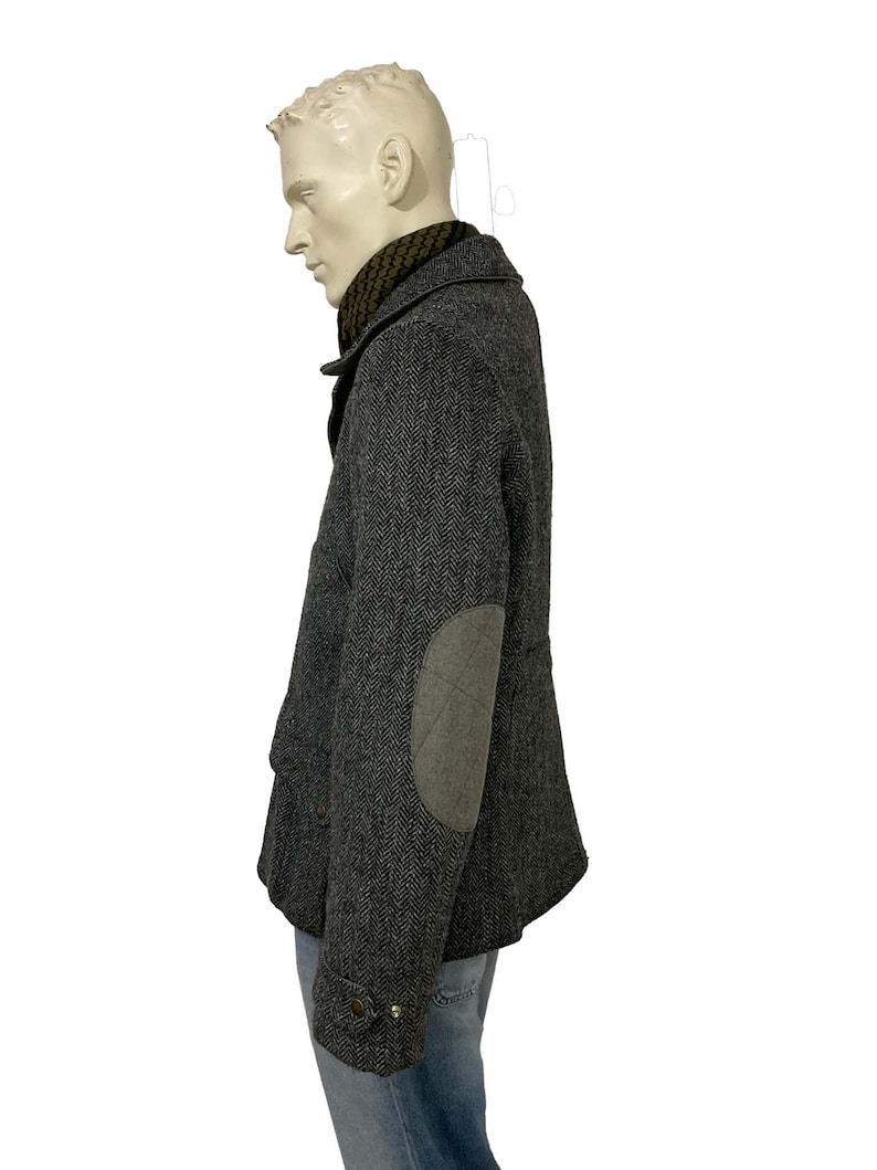 May include: A gray herringbone tweed jacket with elbow patches. The jacket has a notched collar, button closures, and a dark green scarf. The jacket is displayed on a mannequin.