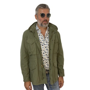 May include: A man wearing a green hooded jacket, a white and blue patterned shirt, blue jeans, and round sunglasses.