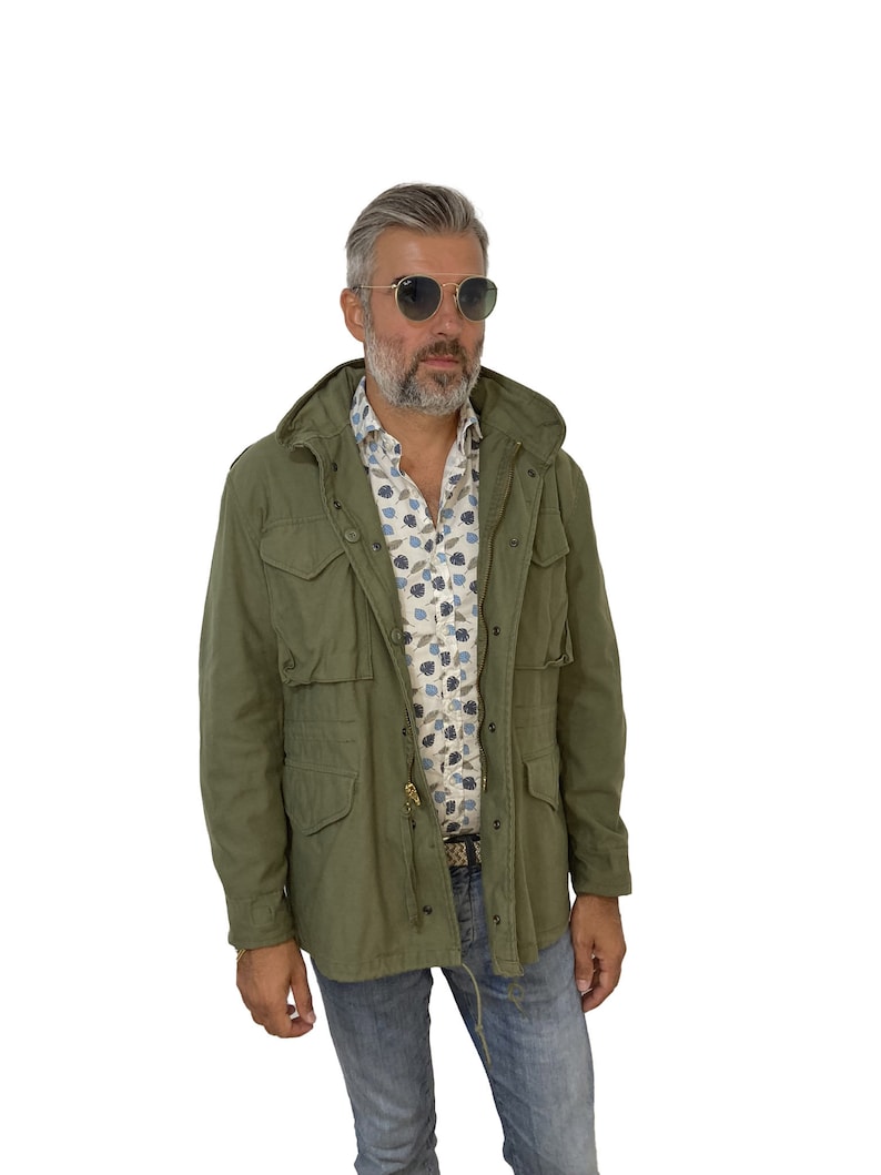 May include: A man wearing a green hooded jacket, a white and blue patterned shirt, blue jeans, and round sunglasses.