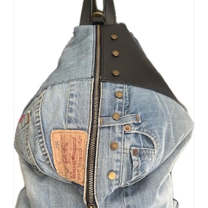 Peut inclure: Un sac à dos bandoulière triangulaire fabriqué en tissu denim bleu clair et en matériau noir texturé. Le sac présente une fermeture éclair verticale centrale, des rivets en métal couleur laiton et une étiquette en cuir beige Levi Strauss & Co. 501.