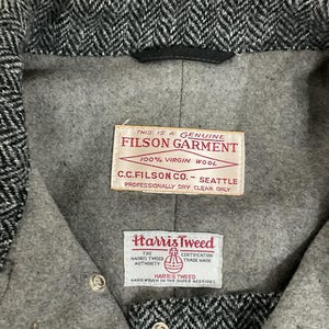 May include: Close-up of a gray and black herringbone tweed jacket interior. The jacket features two labels: "FILSON GARMENT" and "Harris Tweed." The labels provide details about the garment's materials and origin.