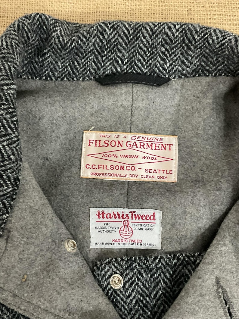 May include: Close-up of a gray and black herringbone tweed jacket interior. The jacket features two labels: "FILSON GARMENT" and "Harris Tweed." The labels provide details about the garment's materials and origin.