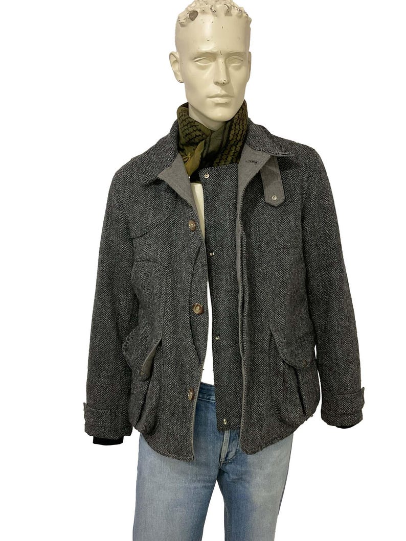 May include: A gray herringbone jacket with a gray collar and pockets, displayed on a mannequin. The jacket has brown buttons and is paired with a green and black scarf and blue jeans.