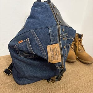 May include: A denim backpack with a black leather top and straps. The backpack features a zipper closure and a Levi Strauss & Co. label. The bag is made from blue denim and has a vintage look. A pair of brown boots are in the background.