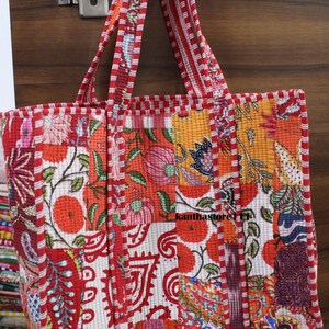 May include: A quilted tote bag with a patchwork design. The bag features a vibrant mix of floral patterns in red, orange, and white. The handles and edges are trimmed with a red and white striped fabric. The bag is hanging on a wooden door.