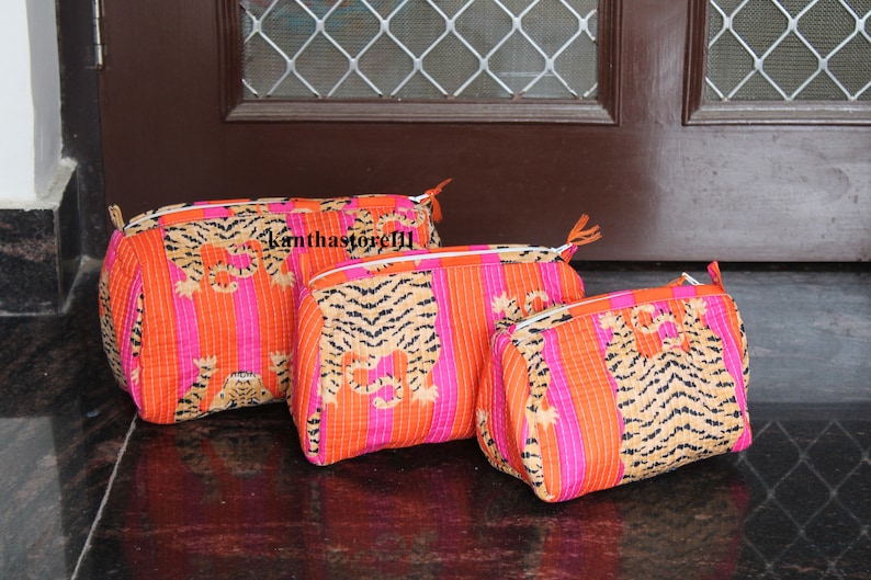 May include: Three pink and orange striped fabric cosmetic bags with a tiger print. The bags are lined up in a row from largest to smallest.