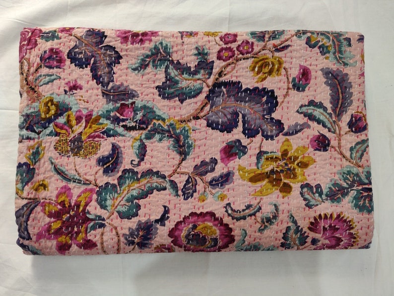Op de afbeelding: Een roze kantha-deken met een bloemenpatroon in tinten paars, blauw en geel. De deken is gemaakt van katoen en heeft een handgestikt ontwerp.
