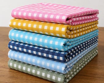 May include: A stack of folded fabric in various colors, including pink, yellow, light blue, navy, and green. Each fabric has a white and colored checkered pattern. The fabrics are neatly stacked on a wooden surface.