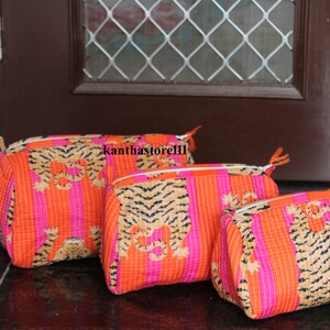 May include: Three quilted makeup bags with a tiger print design. The bags are pink and orange with black stripes and are lined up in order of size, with the largest bag at the back and the smallest bag at the front.