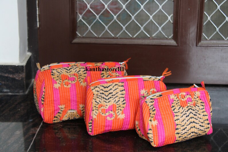 May include: Three quilted makeup bags with a tiger print design. The bags are pink and orange with black stripes and are lined up in order of size, with the largest bag at the back and the smallest bag at the front.