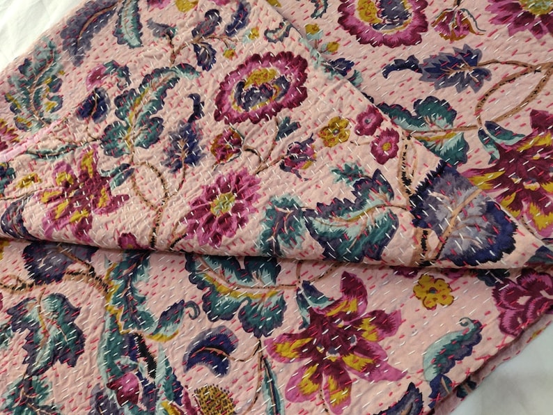 Op de afbeelding: Een roze kantha-deken met een bloemenpatroon in tinten roze, paars, groen en geel. De deken is gemaakt van katoen en heeft een handgestikt ontwerp.