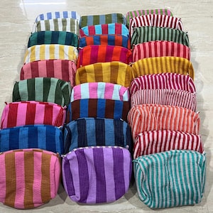 Op de afbeelding: Een verzameling rechthoekige etuis met ritssluiting, gerangschikt in drie kolommen. Elk etui heeft een opvallend verticaal streeppatroon in diverse kleuren zoals roze, blauw, geel, groen, paars, oranje en rood met een textuurrijke afwerking.