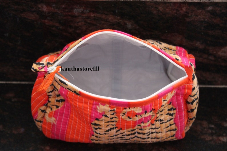 May include: A bright orange, pink and brown quilted makeup bag with a zipper closure and a tassel pull. The bag is open and the inside is lined with a light grey fabric.