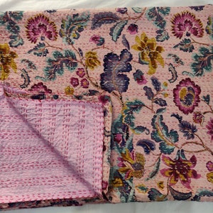 Op de afbeelding: Een roze kantha-deken met een bloemenpatroon in tinten paars, blauw en geel. De deken is doormidden gevouwen, waardoor de roze stiksels aan de achterkant zichtbaar zijn.