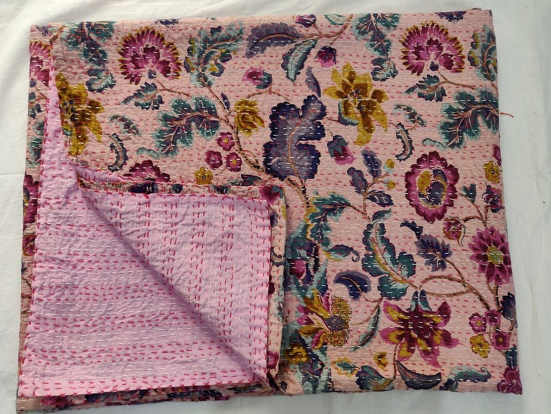 Op de afbeelding: Een roze kantha-deken met een bloemenpatroon in tinten paars, blauw en geel. De deken is doormidden gevouwen, waardoor de roze stiksels aan de achterkant zichtbaar zijn.