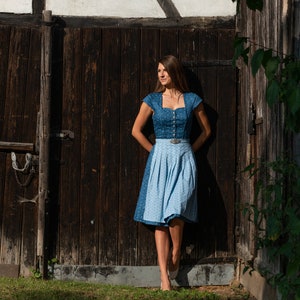Puede incluir: Una mujer con un vestido de dirndl azul y blanco se encuentra de pie frente a una puerta de madera. El vestido tiene un corpi&ntilde;o azul con ribete blanco y una falda blanca con un estampado floral azul. La mujer est&aacute; mirando hacia la izquierda de la imagen.