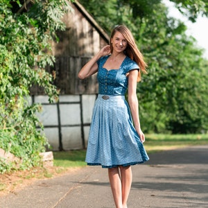 Puede incluir: Una mujer con un vestido de dirndl azul y blanco con un cintur&oacute;n blanco y una hebilla plateada. Est&aacute; de pie en un camino pavimentado con un edificio de madera en el fondo.