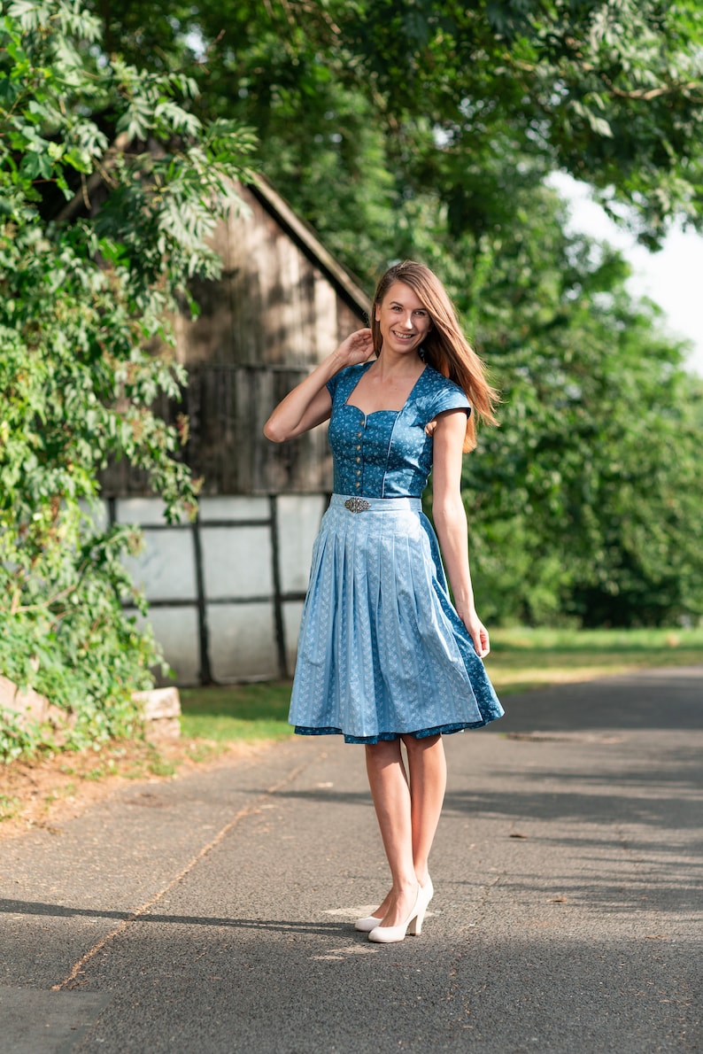 Puede incluir: Una mujer con un vestido de dirndl azul y blanco con un cintur&oacute;n blanco y una hebilla plateada. Est&aacute; de pie en un camino pavimentado con un edificio de madera en el fondo.