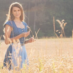 Puede incluir: Una mujer con un vestido estampado en azul y blanco se encuentra de pie en un campo de hierba alta. Est&aacute; sosteniendo algunos tallos de hierba en su mano y sonriendo a la c&aacute;mara.