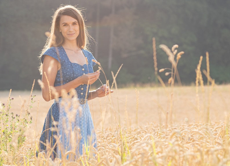 Puede incluir: Una mujer con un vestido estampado en azul y blanco se encuentra de pie en un campo de hierba alta. Est&aacute; sosteniendo algunos tallos de hierba en su mano y sonriendo a la c&aacute;mara.