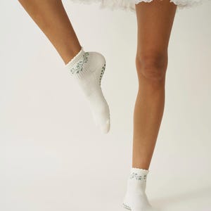 May include: White ankle socks featuring a light blue floral pattern at the cuff. The socks have a ribbed design and small, dark grey grips on the soles. The image shows the socks being worn.