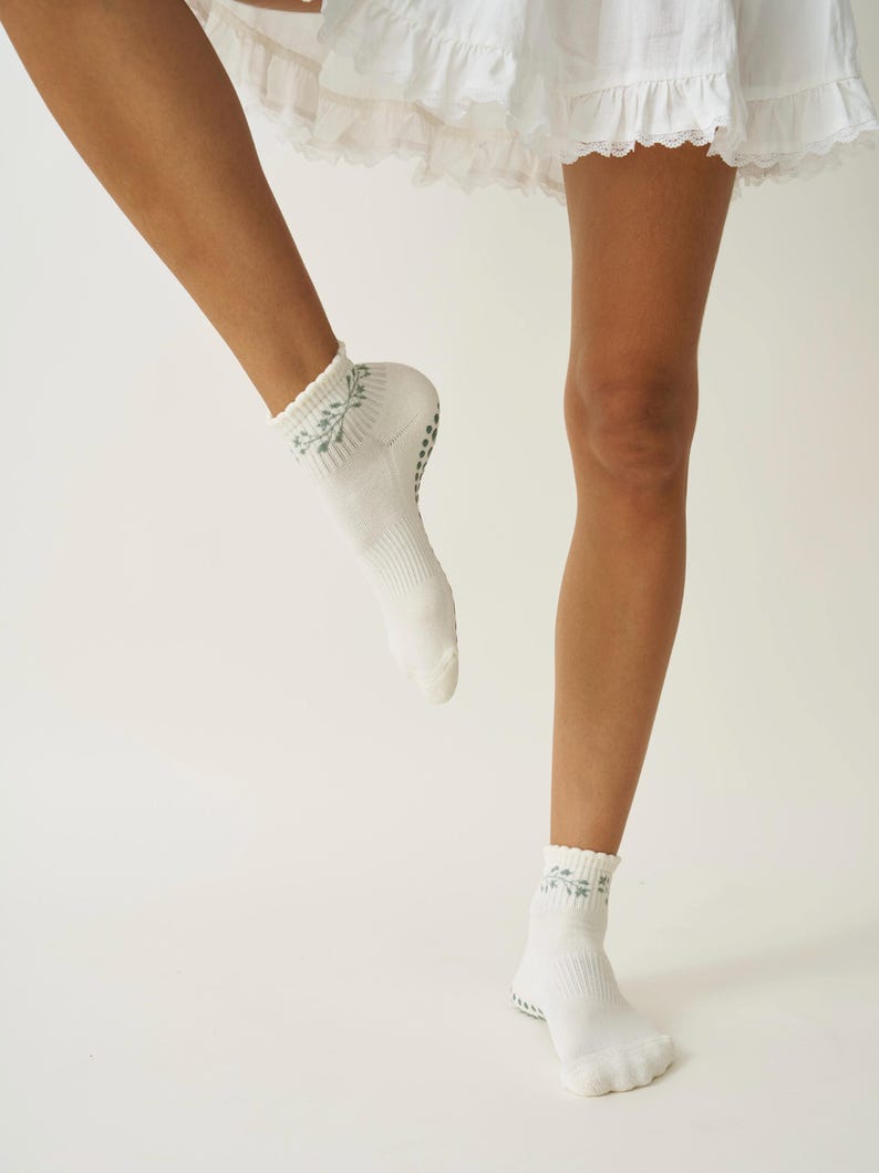 May include: White ankle socks featuring a light blue floral pattern at the cuff. The socks have a ribbed design and small, dark grey grips on the soles. The image shows the socks being worn.