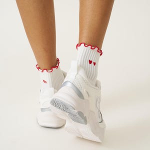 May include: White athletic shoes with silver accents and white socks. The socks have a red ruffled trim and two small red hearts. The shoes are chunky and have a textured sole.