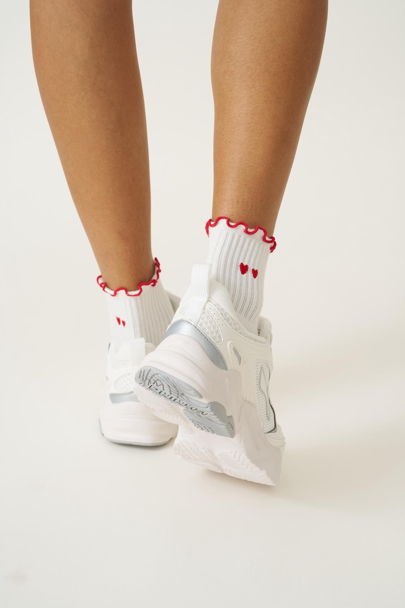 May include: White athletic shoes with silver accents and white socks. The socks have a red ruffled trim and two small red hearts. The shoes are chunky and have a textured sole.