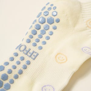 May include: Close-up of a cream-colored sock with blue non-slip grips in a hexagonal pattern. The word "EFFOI" is printed on the side. Smiley faces in yellow and purple are printed on the sock.
