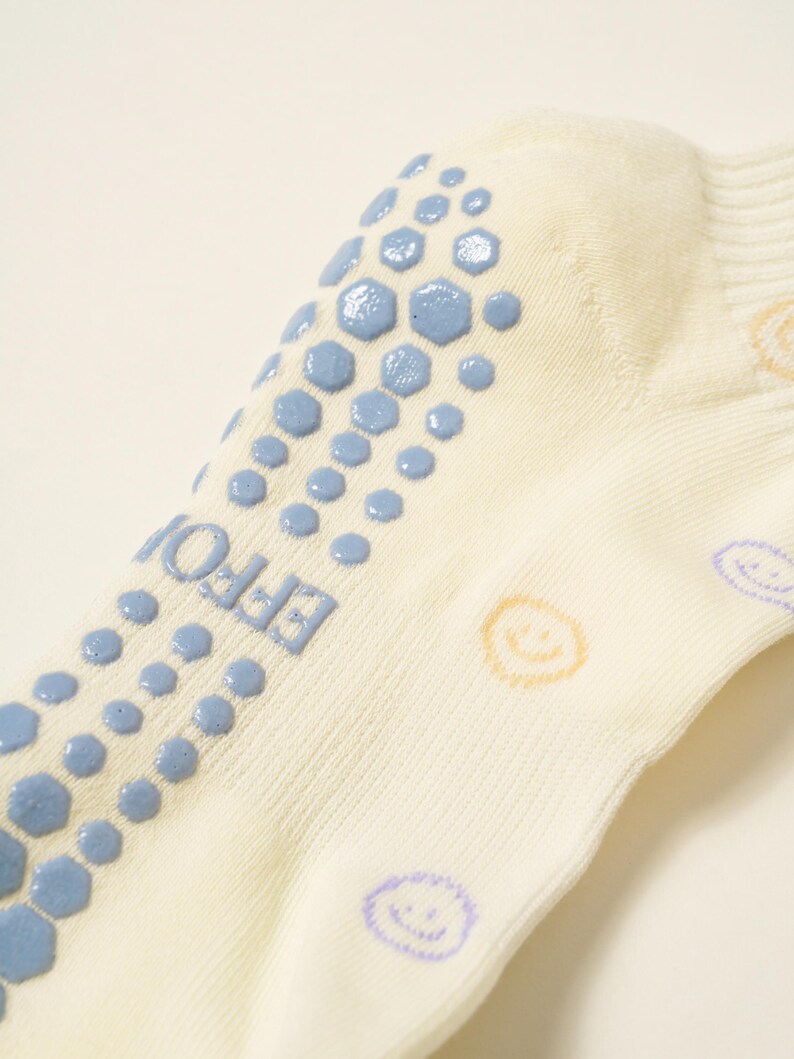 May include: Close-up of a cream-colored sock with blue non-slip grips in a hexagonal pattern. The word "EFFOI" is printed on the side. Smiley faces in yellow and purple are printed on the sock.