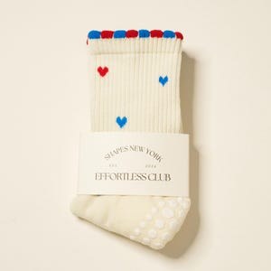 Könnte beinhalten: Cremefarbene Socken mit roten und blauen Verzierungen und kleinen Herzmustern. Die Socken haben eine rutschfeste Sohle und ein Etikett mit der Aufschrift "SHAPES NEW YORK EST 2024 EFFORTLESS CLUB."