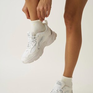 May include: White athletic sneakers with a chunky sole and mesh detailing. The sneakers have a lace-up closure and are paired with white ribbed socks. The shoes are a modern style, suitable for athletic activities or casual wear.